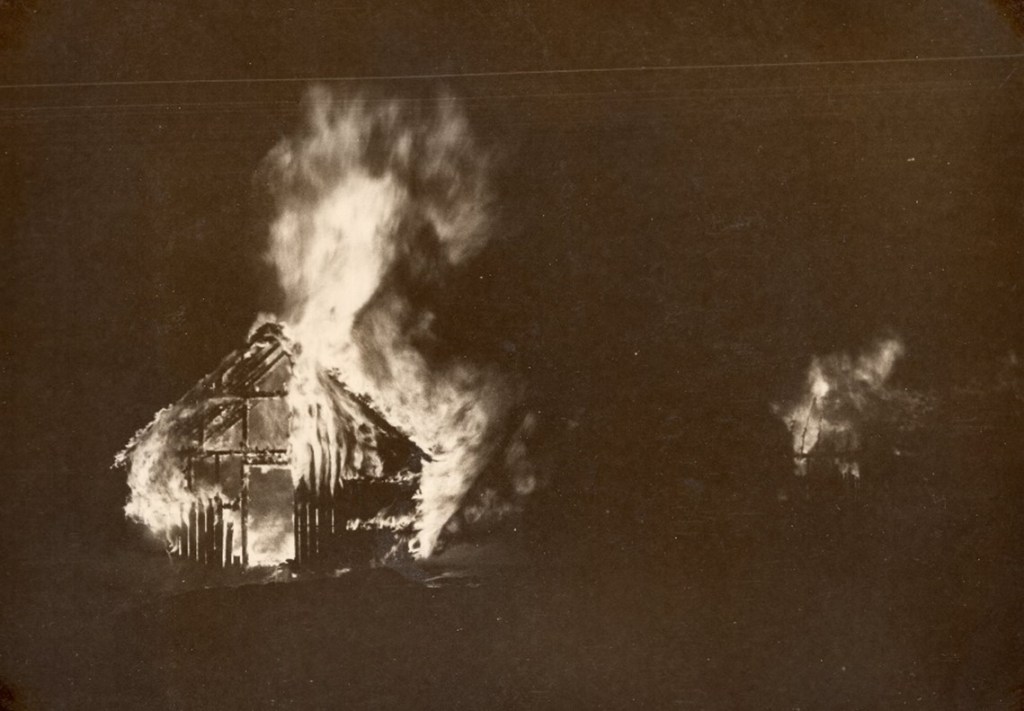 Black and white image of a building on fire.  On the left side, the building is lit up by fire shooting out of the top and sides.  The wood framing is exposed, and the siding is almost completely burnt.  In the background on the right, another fire is seen at the other end of the building. 